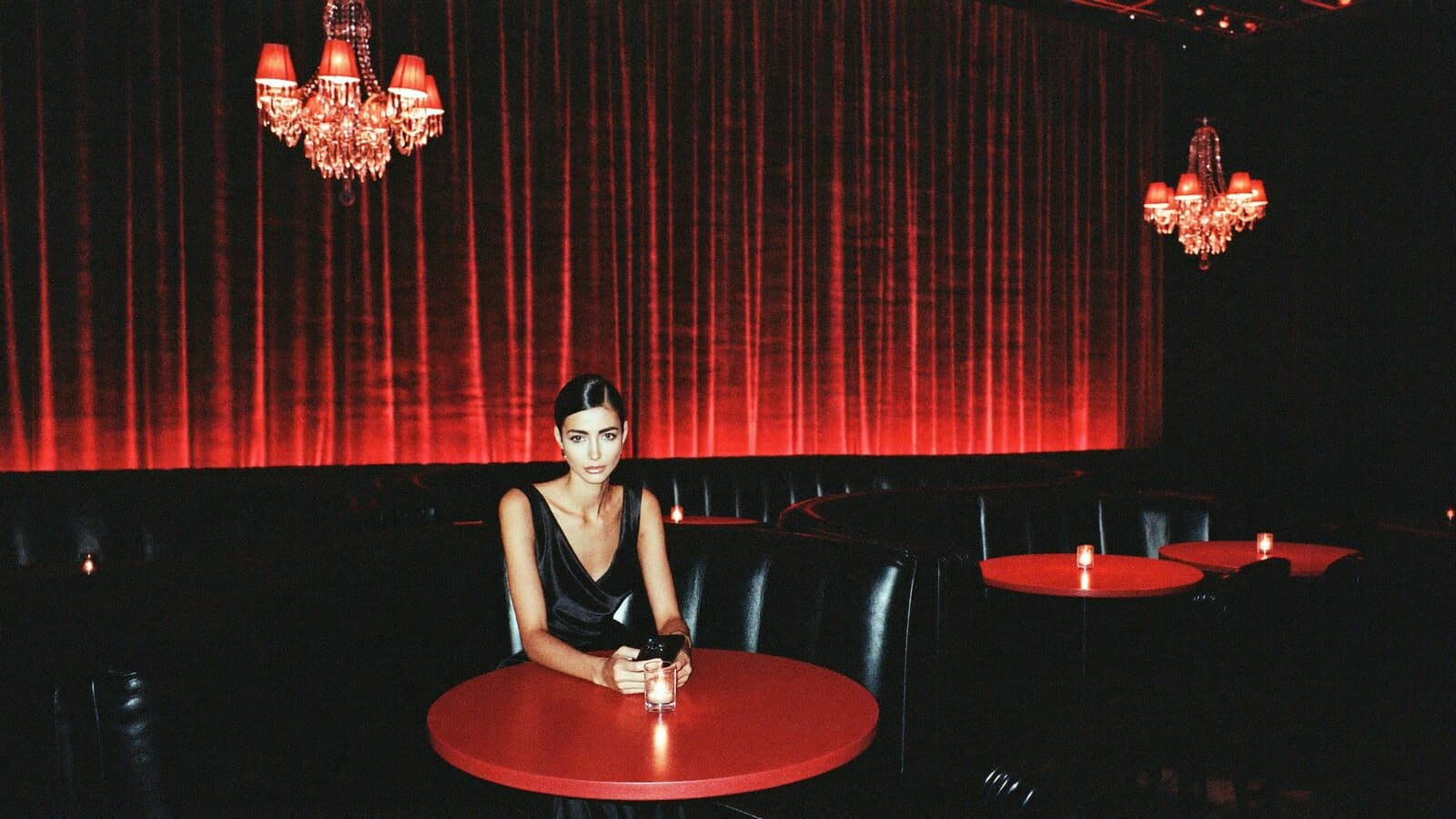 A woman in a black dress sitting alone at a red table lit by a candle in a dark velvet room