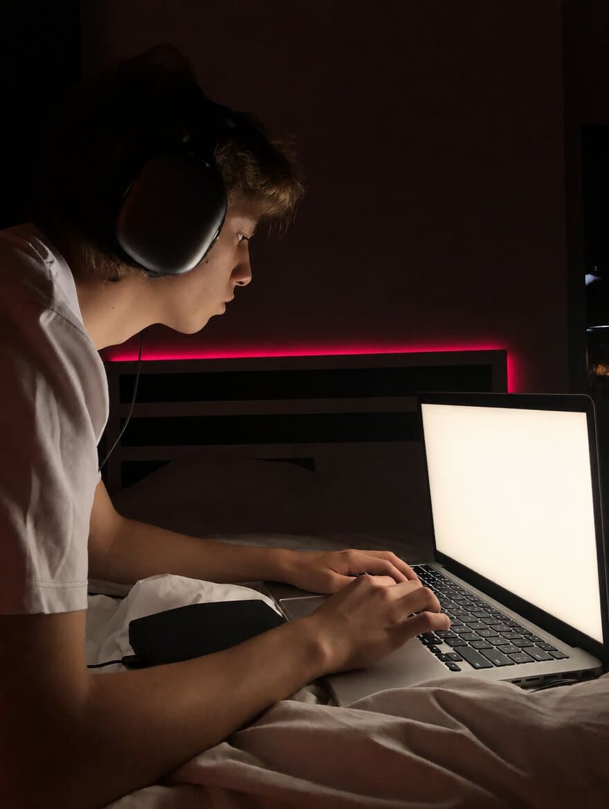 A young man in headphones typing on a laptop in a dark, pink-lit bedroom
