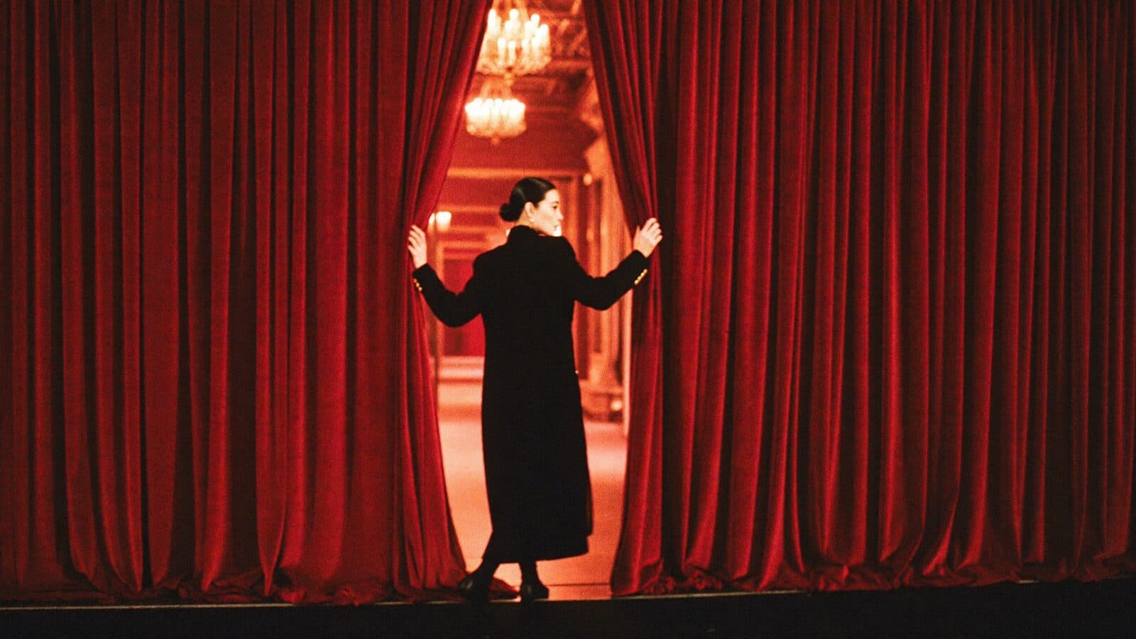 A woman in a long black coat parting red velvet curtains and stepping through into a lit hallway