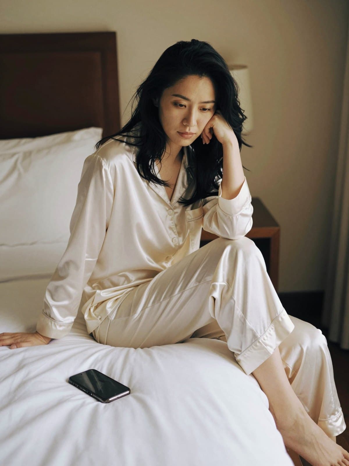 A creator in cream silk pyjamas on the edge of a hotel bed, her phone face down on the sheets