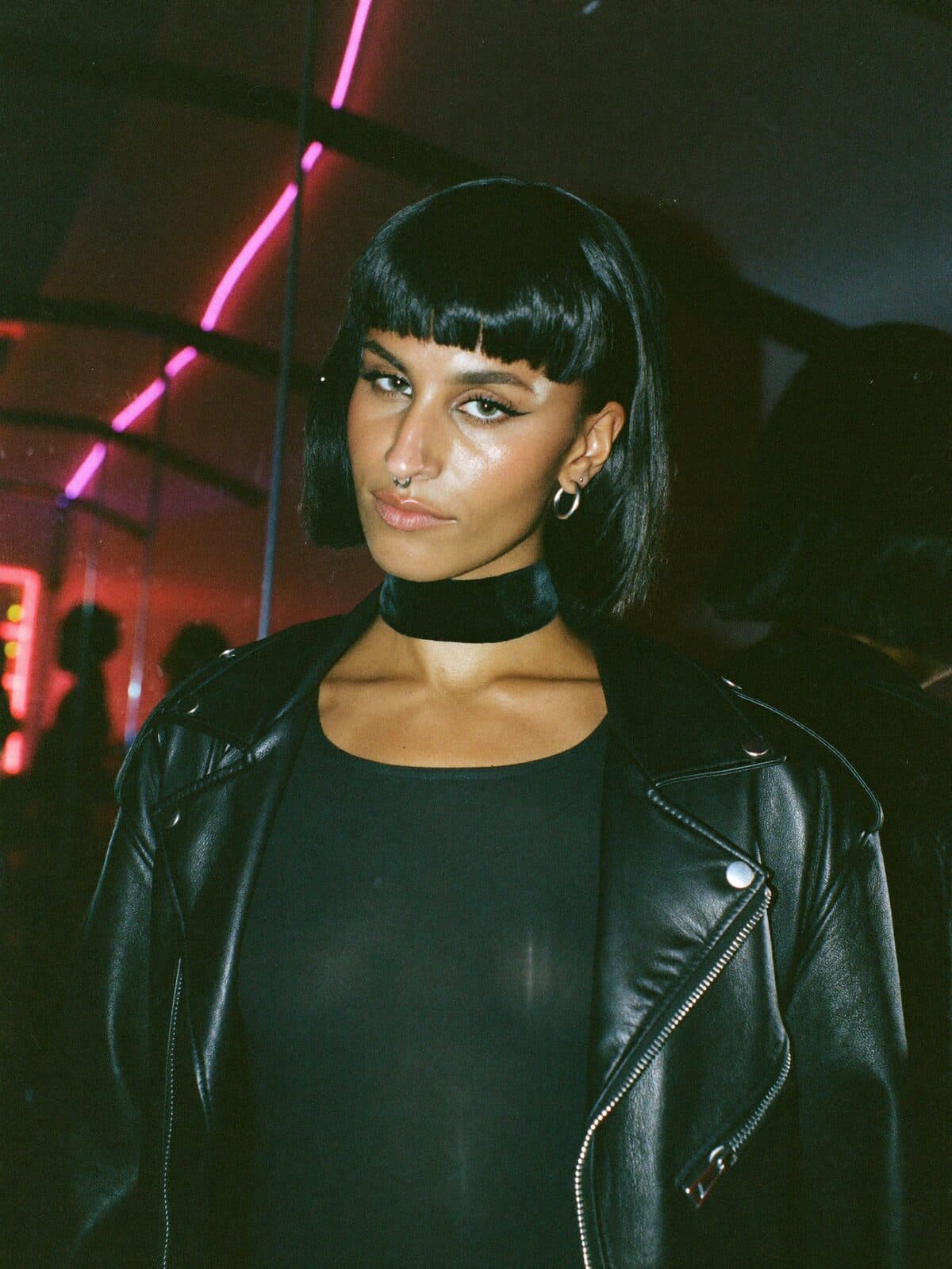 A woman with a black bob and a leather jacket looking straight into the camera in a neon-lit club