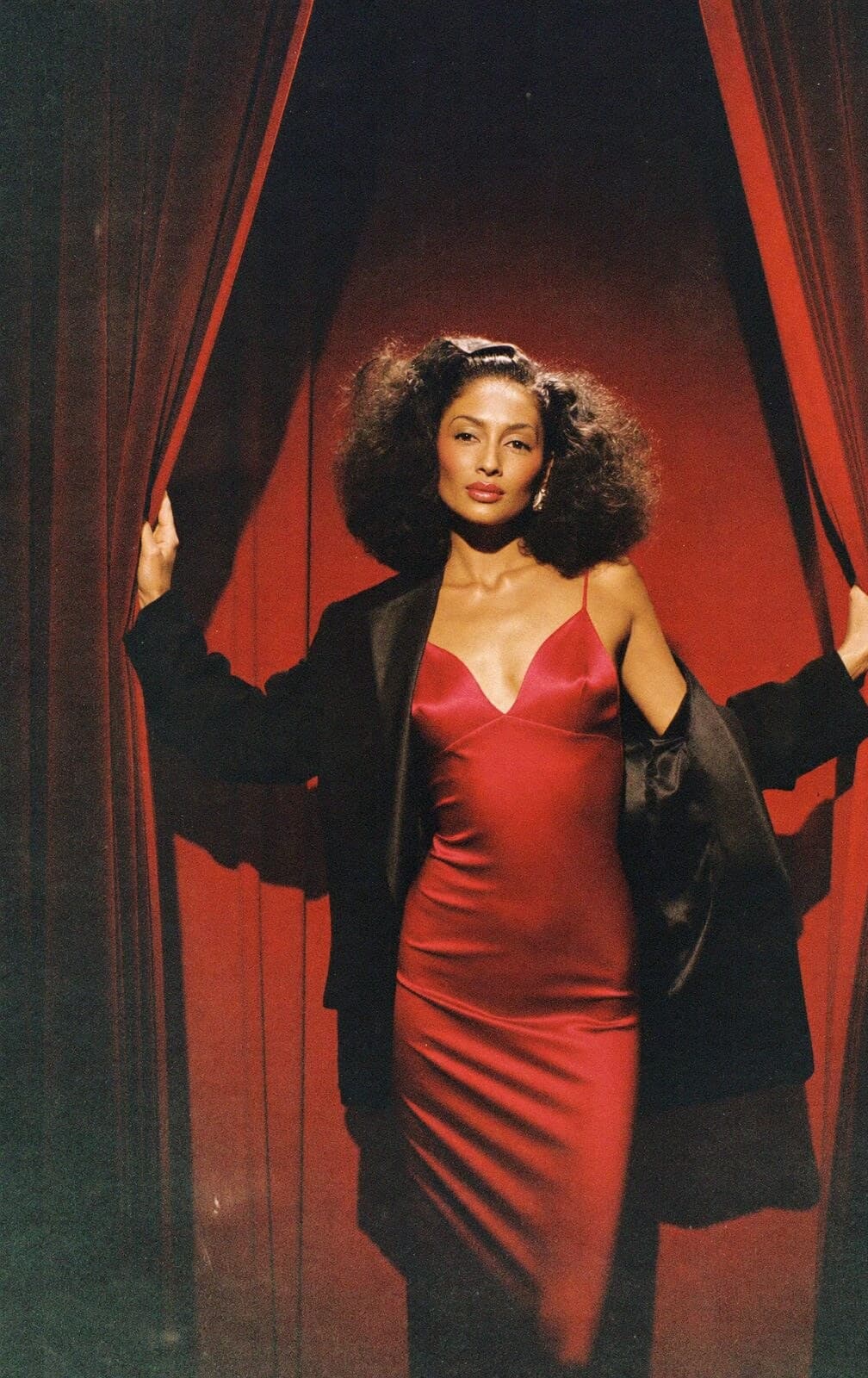 A woman in a red satin slip dress and a black jacket stepping through red curtains into a spotlight
